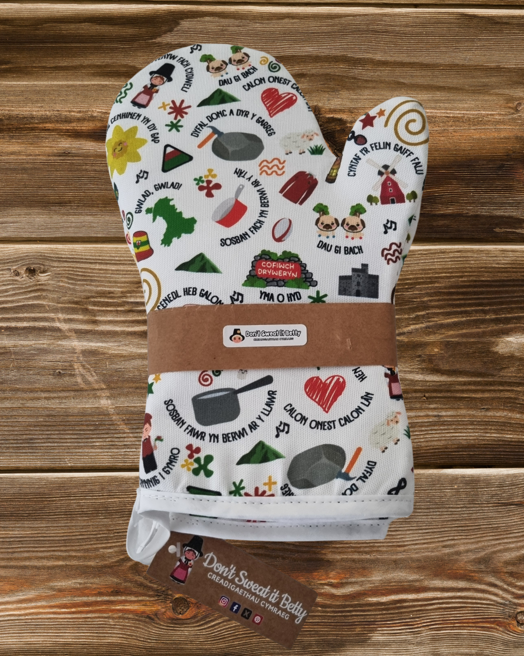 A white oven glove with various colorful Welsh-themed illustrations and text, such as leeks, sheep, and Welsh proverbs on a wooden surface with branding.