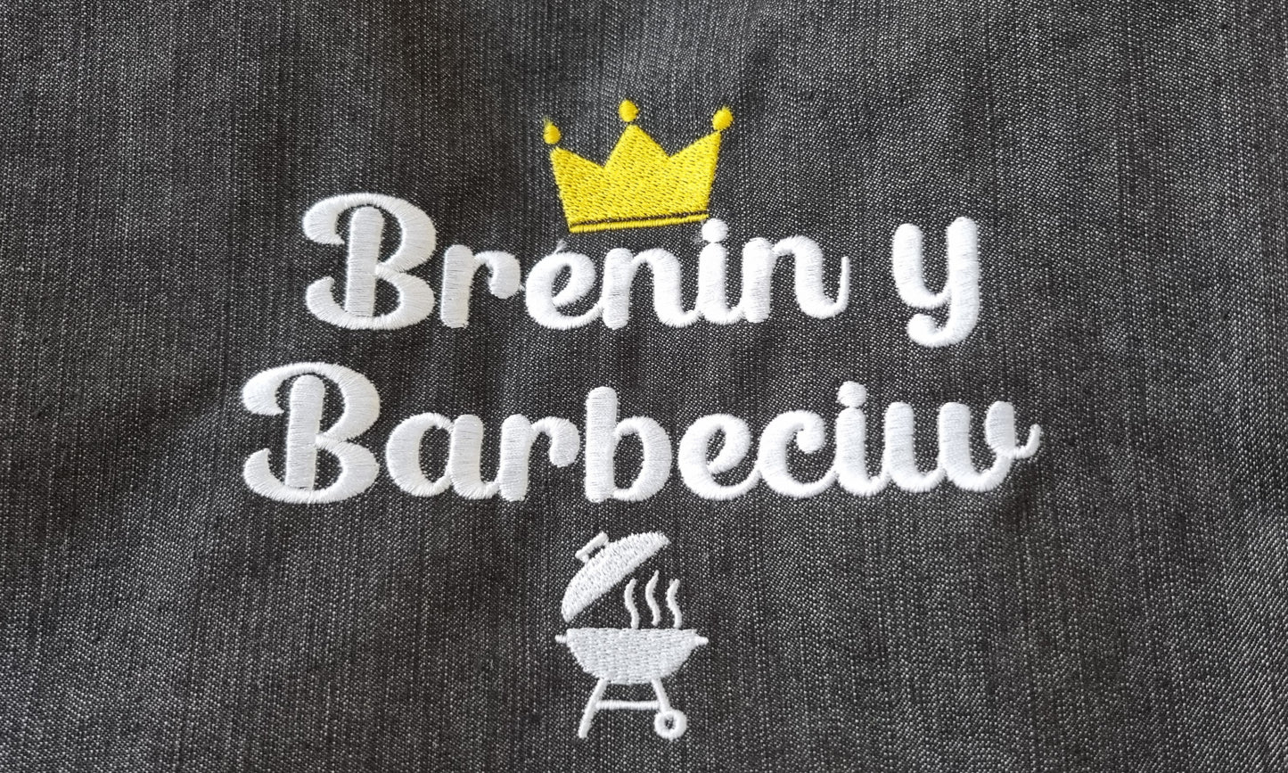 Close-up of an apron with 'Brenin y Barbeciw' embroidery, featuring a crown and a small illustration of a barbecue grill.