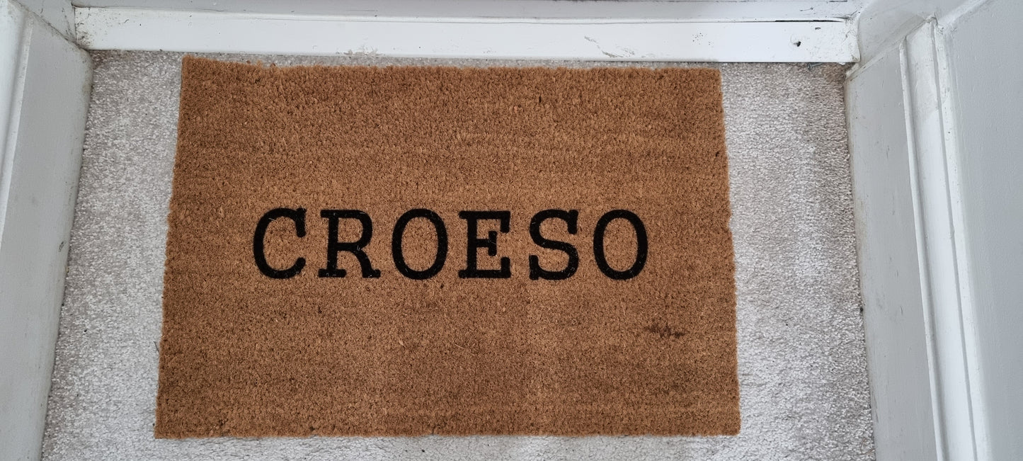A rectangular coir doormat with the Welsh word 'CROESO' printed in the center, placed on a doorstep.