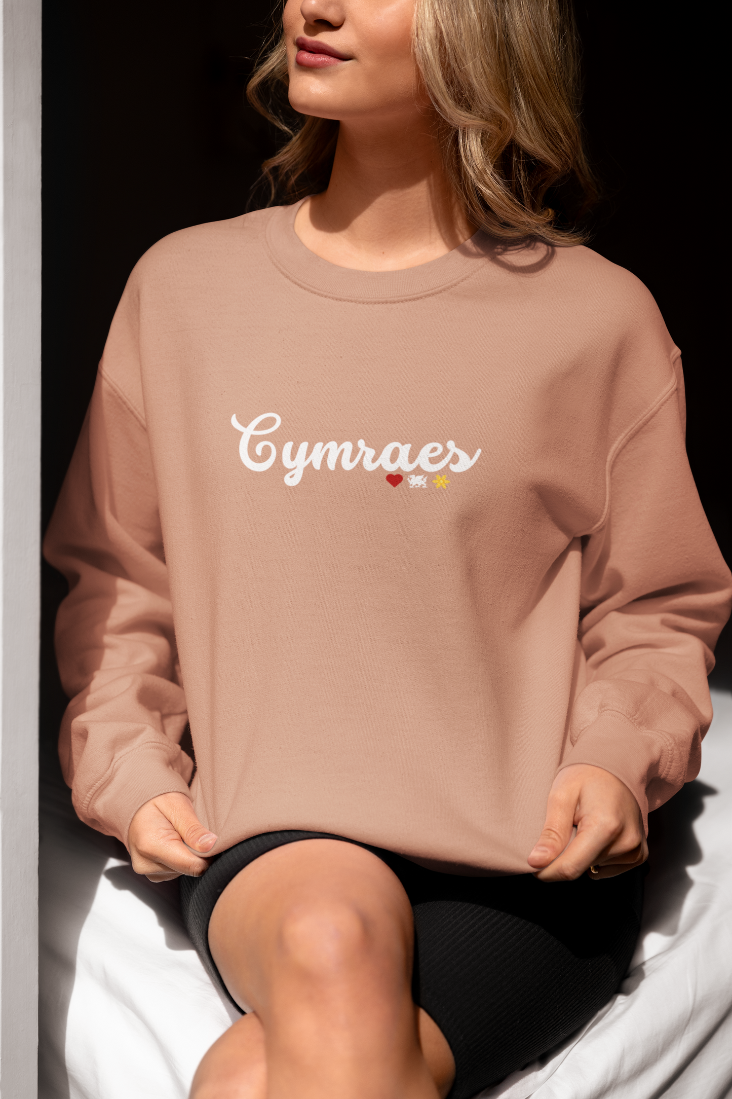 A woman sitting and wearing a pink sweatshirt with the word 'Cymraes' embroidered on the front in white.