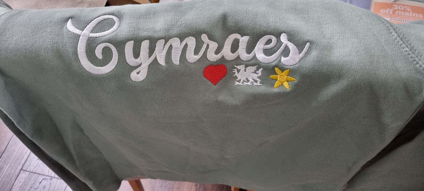 Green fabric with embroidered text and symbols, including 'Cymraes', a heart, a dragon, and a flower.