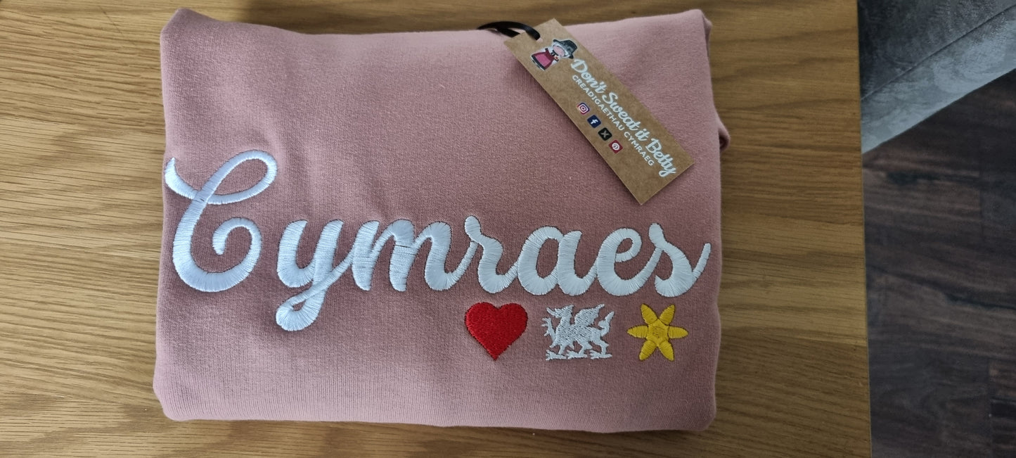 Pink sweater with 'Cymraes' text and Welsh symbols on a wooden surface