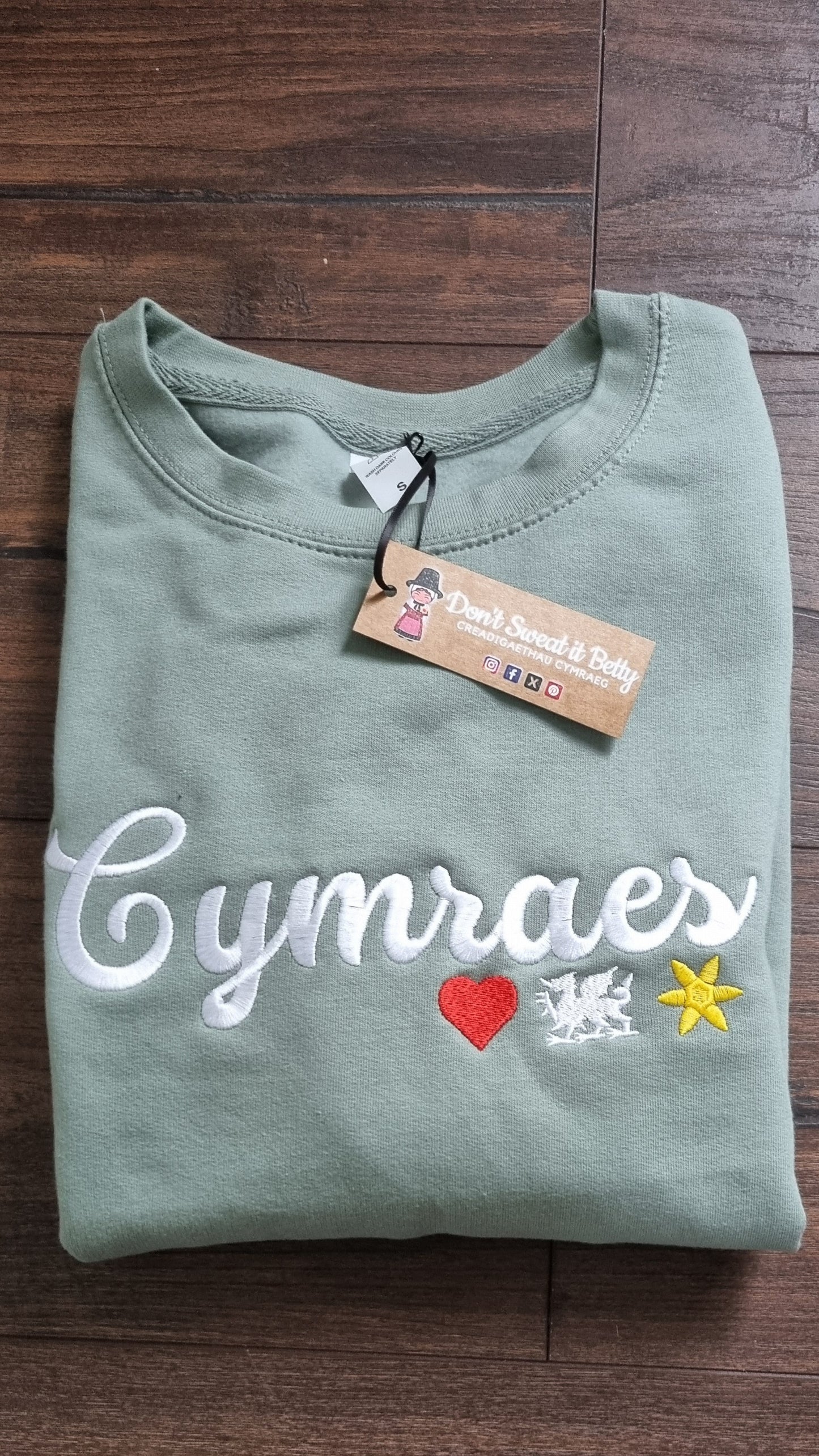 Folded green shirt with 'Cymraes' text and colorful accents on a wooden surface