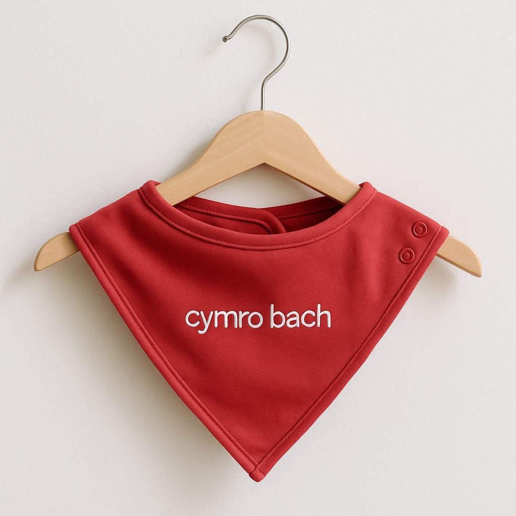 A red embroidered baby bib with 'cymro bach' displayed on a wooden hanger.