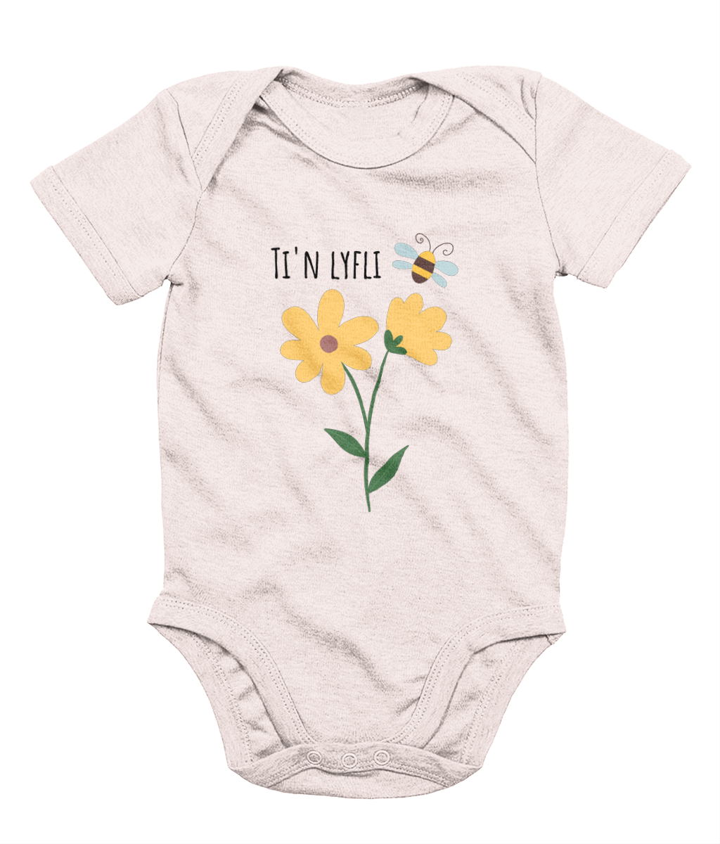 A pink babygrow with a graphic print of a sunflower and a bee, and the Welsh phrase 'Ti'n Lyfli' written in the center.