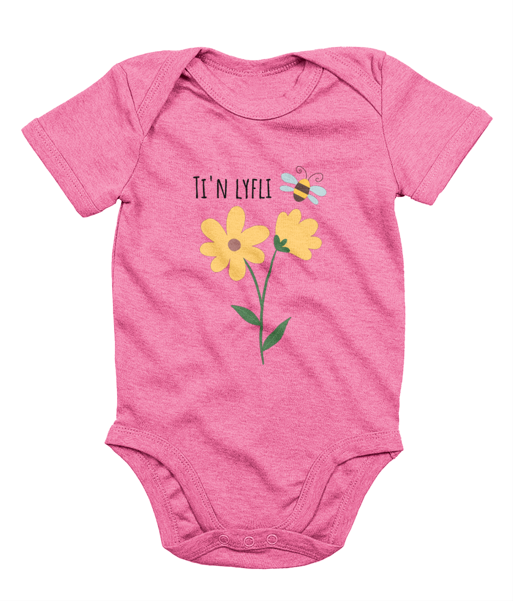 A pink babygrow with a graphic print of a sunflower and a bee, and the Welsh phrase 'Ti'n Lyfli' written in the center.