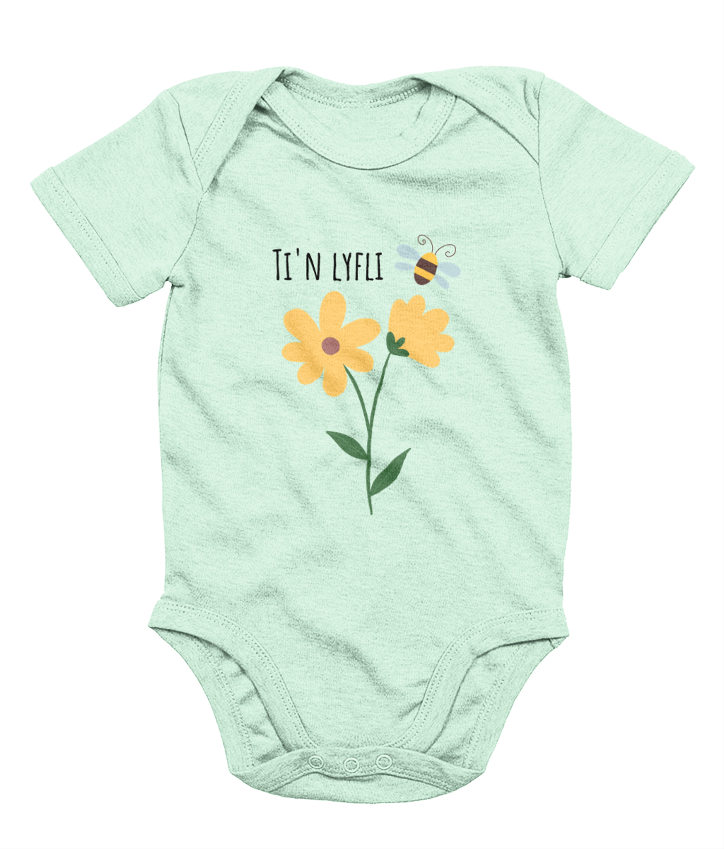 A green babygrow with a graphic print of a sunflower and a bee, and the Welsh phrase 'Ti'n Lyfli' written in the center.