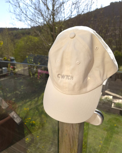 Beige cap with 'CWTCH' branding on a garden wall