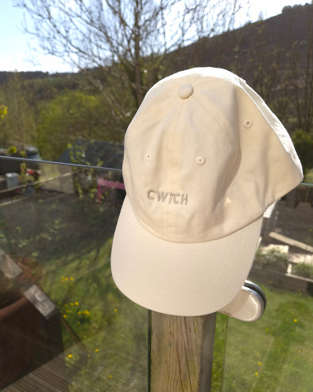Beige cap with 'CWTCH' branding on a garden wall