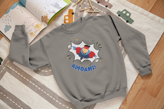 Children's sweatshirt in gray with a colorful front graphic print featuring a superhero character and the Welsh word 'amdani'. The sweatshirt is laid out flat on a wooden surface with children's toys around it.