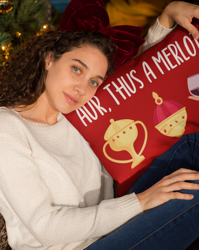 A person holding a red Christmas pillow with the text 'Aur, Thus & Merlot' printed in white, meaning 'Gold, Frankincense and Merlot' in Welsh.