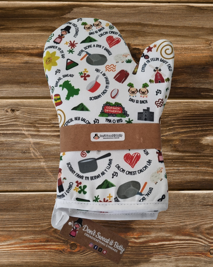 A white oven glove with various colorful Welsh-themed illustrations and text, such as leeks, sheep, and Welsh proverbs on a wooden surface with branding.
