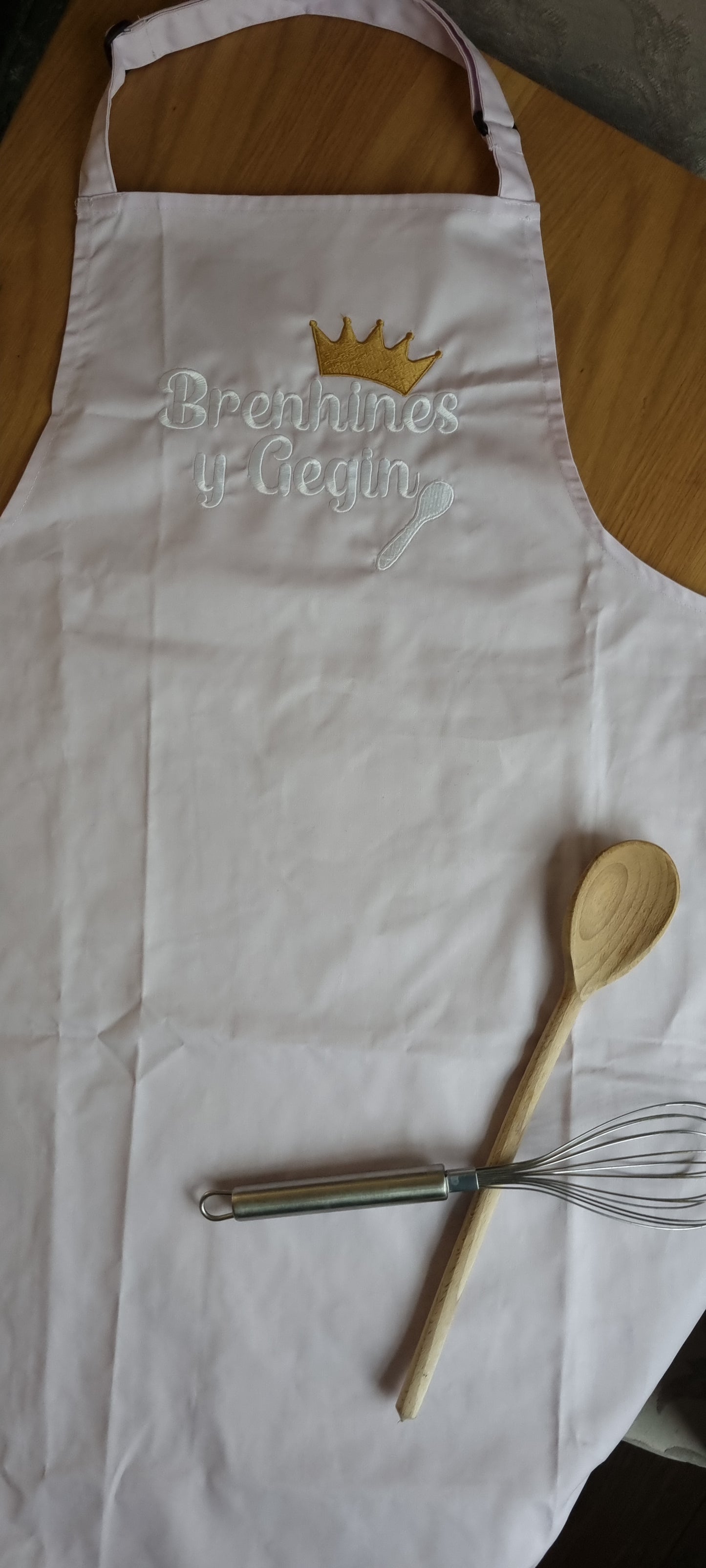 A lilac apron with the embroidered text 'Brenhines y Gegin' displayed at the center, above a wooden spoon and fork.