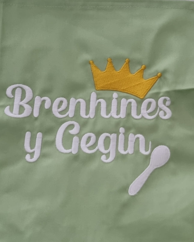 Green apron with embroidered text and crown design on white fabric.