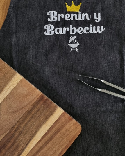 A black adult apron with 'Brenin y Barbeciw' embroidered in white, meaning 'King of the Barbecue', with a crown symbol above the text.