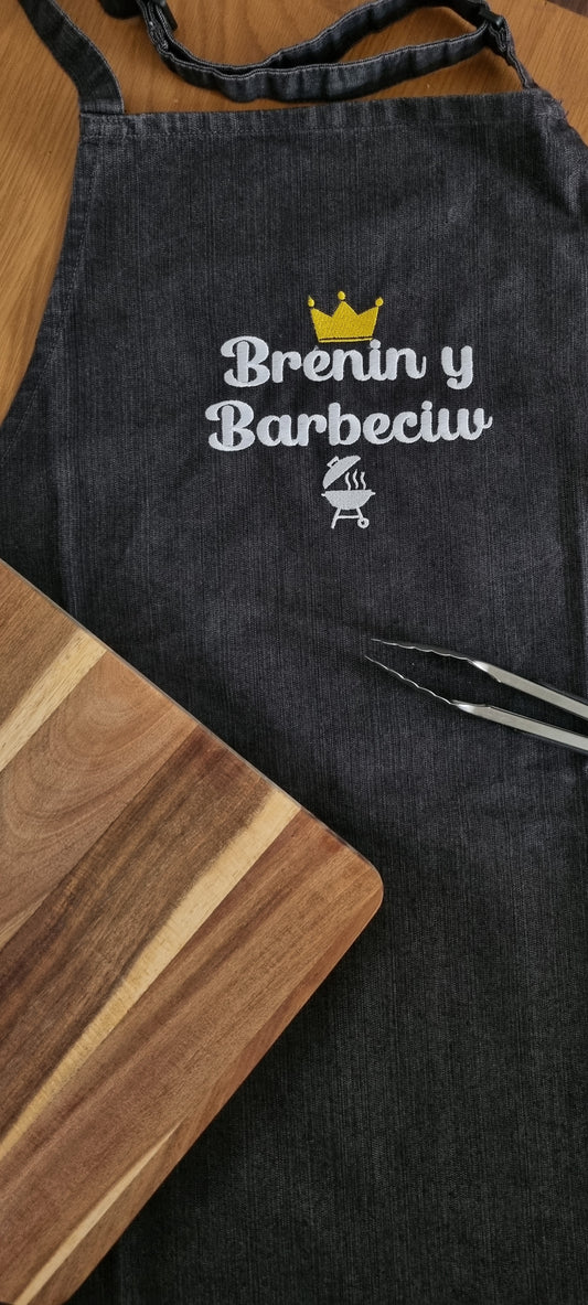 A black adult apron with 'Brenin y Barbeciw' embroidered in white, meaning 'King of the Barbecue', with a crown symbol above the text.
