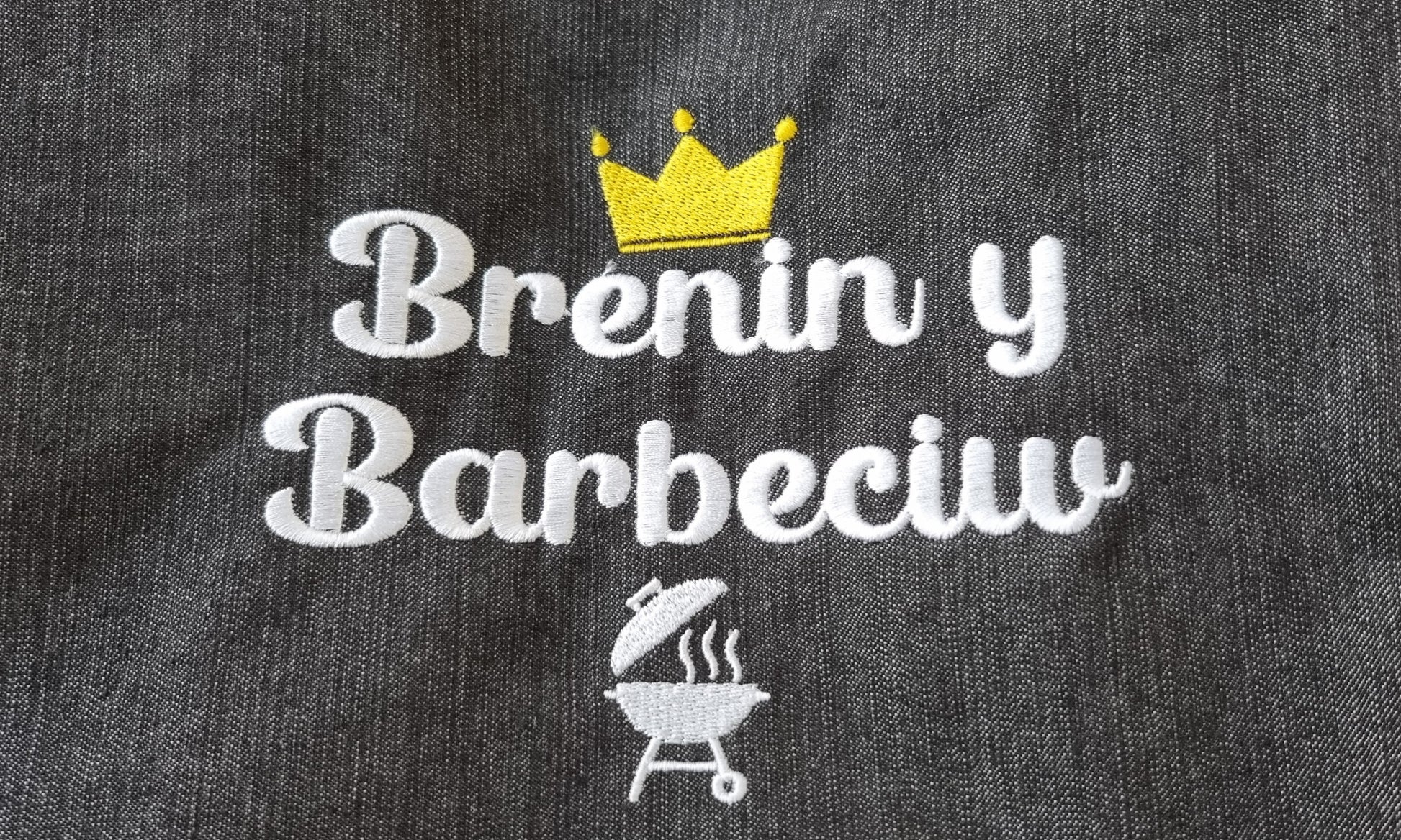 Close-up of an apron with 'Brenin y Barbeciw' embroidery, featuring a crown and a small illustration of a barbecue grill.