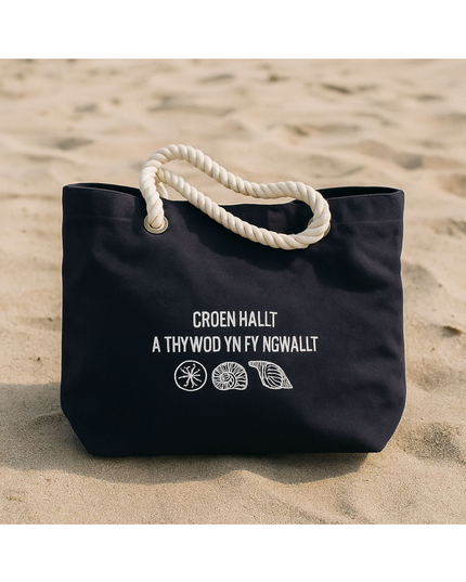 A navy blue beach tote with the embroidered text 'Croen Hallt a Thywod yn fy Ngwallt' in Welsh, featuring a nautical rope handle.