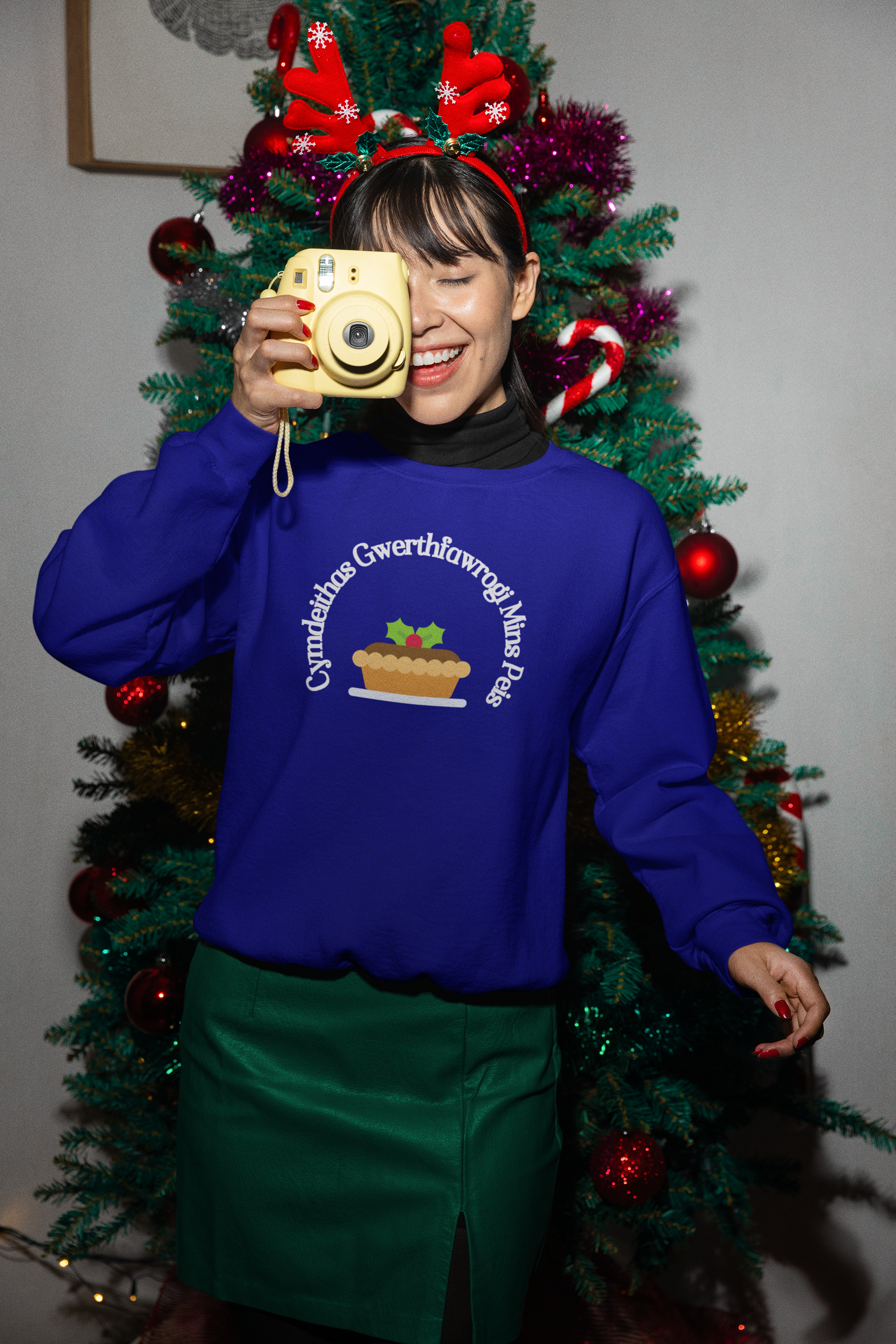 A person wearing a blue sweatshirt with the printed text 'Cymdeithas Gwerthfawrogi Mins Peis' standing in front of a Christmas tree.