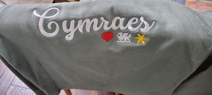 Green fabric with embroidered text and symbols, including 'Cymraes', a heart, a dragon, and a flower.