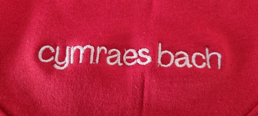Red fabric with embroidered white text 'cymraes bach'.