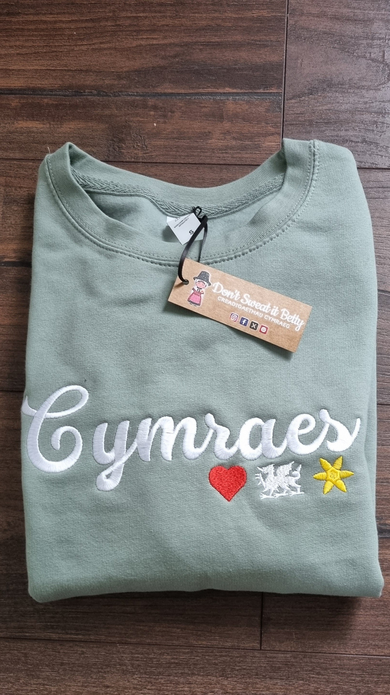 Folded green shirt with 'Cymraes' text and colorful accents on a wooden surface