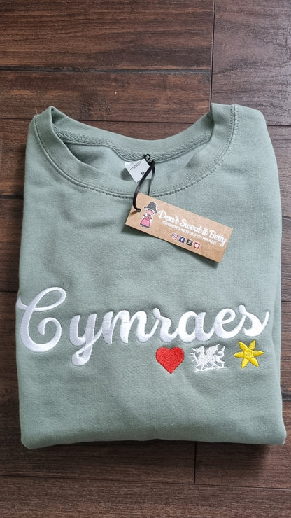 Folded green shirt with 'Cymraes' text and colorful accents on a wooden surface