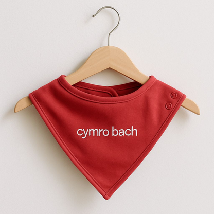 A red embroidered baby bib with 'cymro bach' displayed on a wooden hanger.
