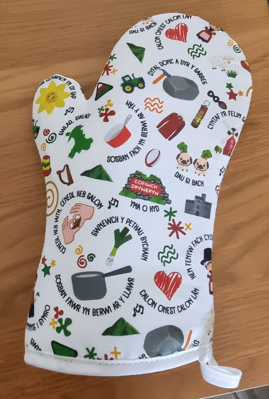 A white oven glove with various colorful Welsh-themed illustrations and text, such as leeks, sheep, and Welsh proverbs.