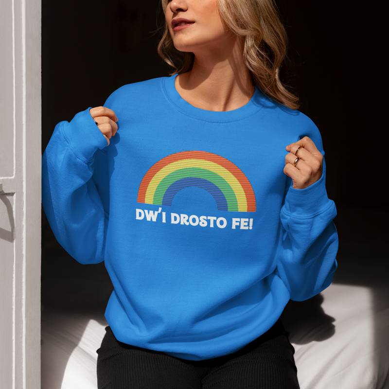 A person wearing a blue sweatshirt with the text 'DWI DROSTO FE' in bold Welsh lettering, against a rainbow background.