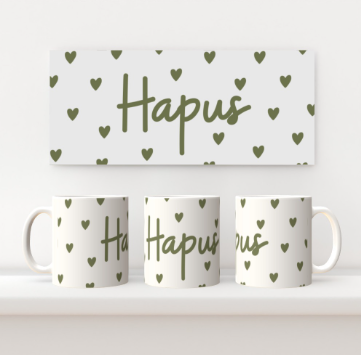 A white ceramic mug with the Welsh word 'Hapus' written on it, accompanied by a pattern of green hearts. The mug is displayed alongside a matching rectangular card with the same design.