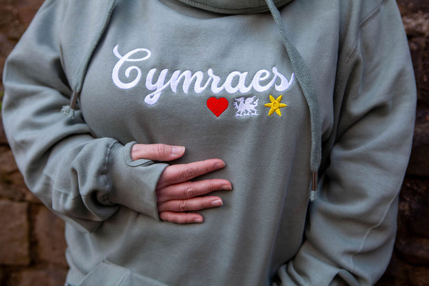 Person wearing a hoodie with 'Cymraes' and symbols on a brick wall background