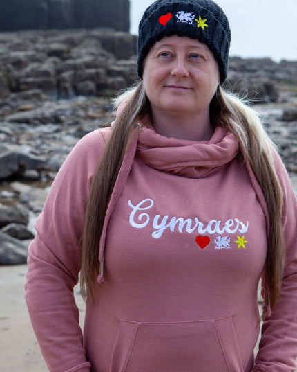 Person wearing a pink hoodie with 'Cymraes' text on a beach