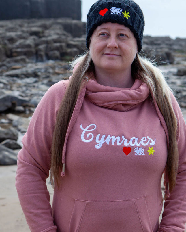 Person wearing a pink hoodie with 'Cymraes' text on a beach