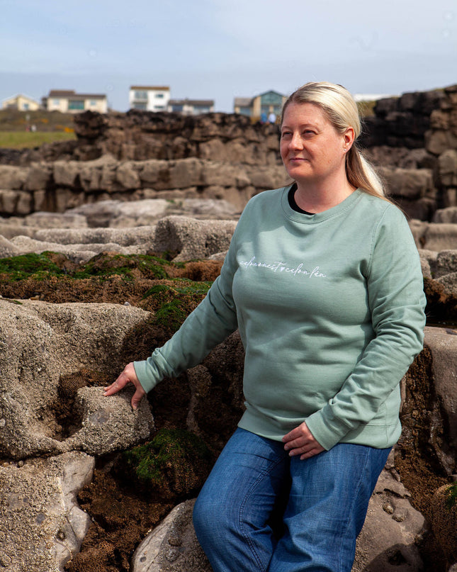 Woman wearing a green sweatshirt with text, standing on stone steps with a scenic background.