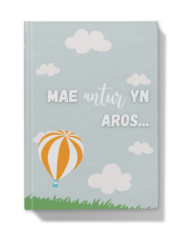 A hardback journal with a light blue cover featuring Welsh text ' Mae Antur yn Aros ' with a graphic of a hot air balloon, clouds, and grass at the bottom.