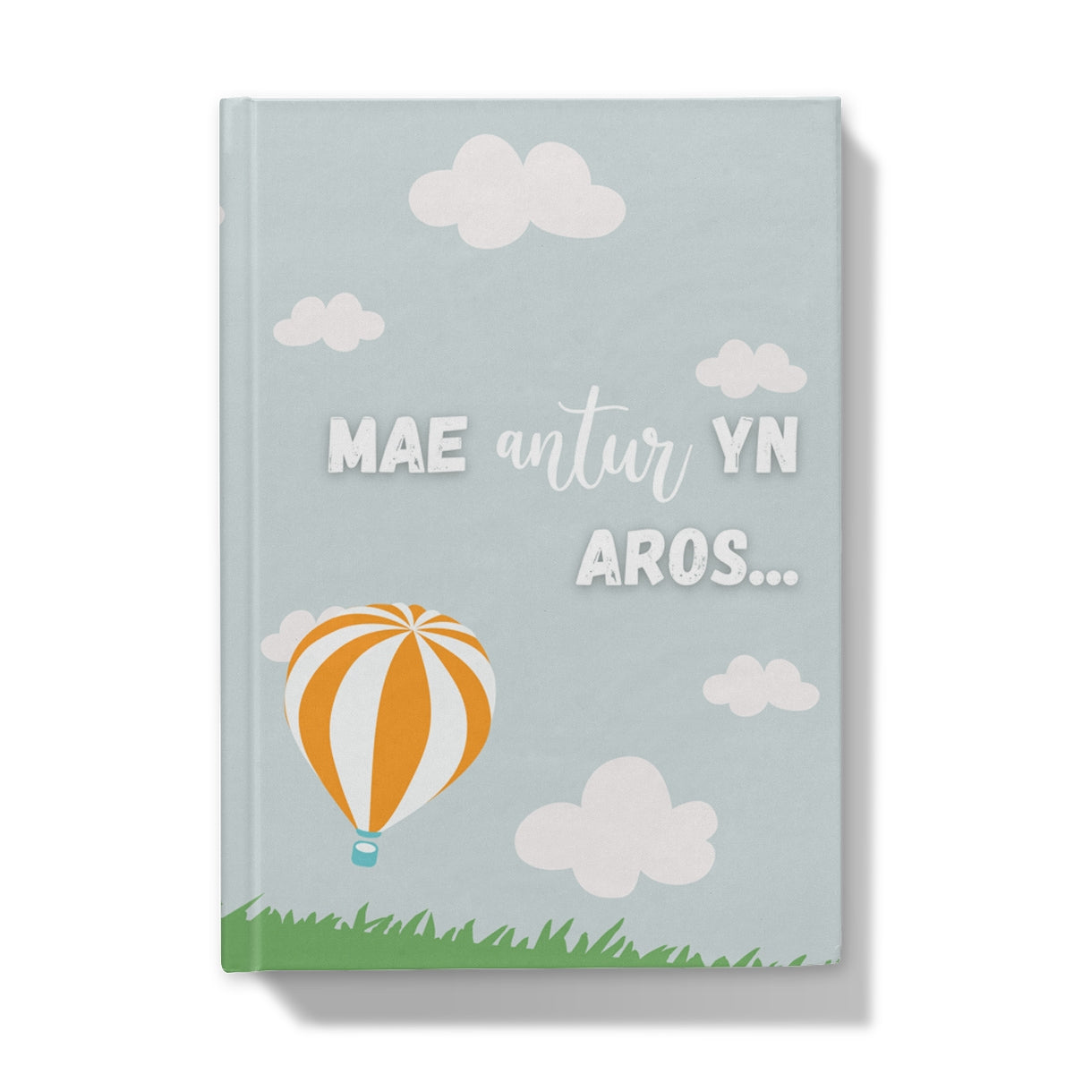 A hardback journal with a light blue cover featuring Welsh text ' Mae Antur yn Aros ' with a graphic of a hot air balloon, clouds, and grass at the bottom.