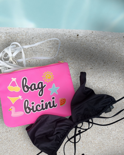 A pink waterproof swim bag with the text 'bag bicini' printed on it, placed on a stone surface near a pool, with a black bikini bottom beside it.