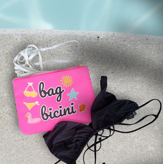 A pink waterproof swim bag with the text 'bag bicini' printed on it, placed on a stone surface near a pool, with a black bikini bottom beside it.