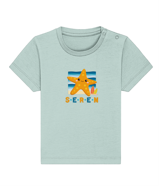 A baby t-shirt in a light color with the name 'Seren' printed on the front, featuring a graphic of a starfish and a sunset or sunrise scene.