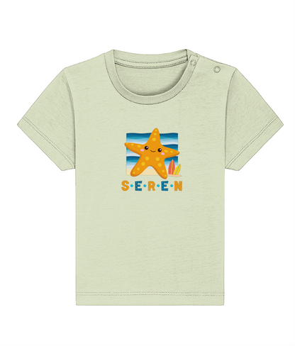 A baby t-shirt in a light color with the name 'Seren' printed on the front, featuring a graphic of a starfish and a sunset or sunrise scene.