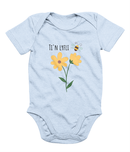 A blue babygrow with a graphic print of a sunflower and a bee, and the Welsh phrase 'Ti'n Lyfli' written in the center.