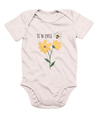 A pink babygrow with a graphic print of a sunflower and a bee, and the Welsh phrase 'Ti'n Lyfli' written in the center.