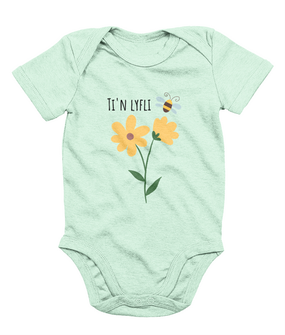 A green babygrow with a graphic print of a sunflower and a bee, and the Welsh phrase 'Ti'n Lyfli' written in the center.