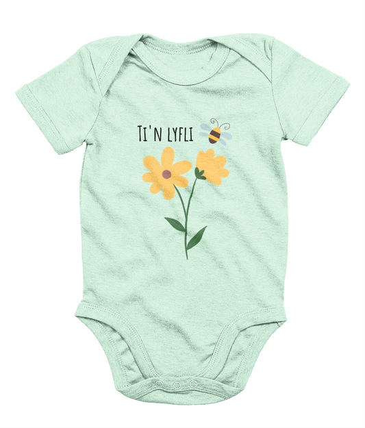 A green babygrow with a graphic print of a sunflower and a bee, and the Welsh phrase 'Ti'n Lyfli' written in the center.