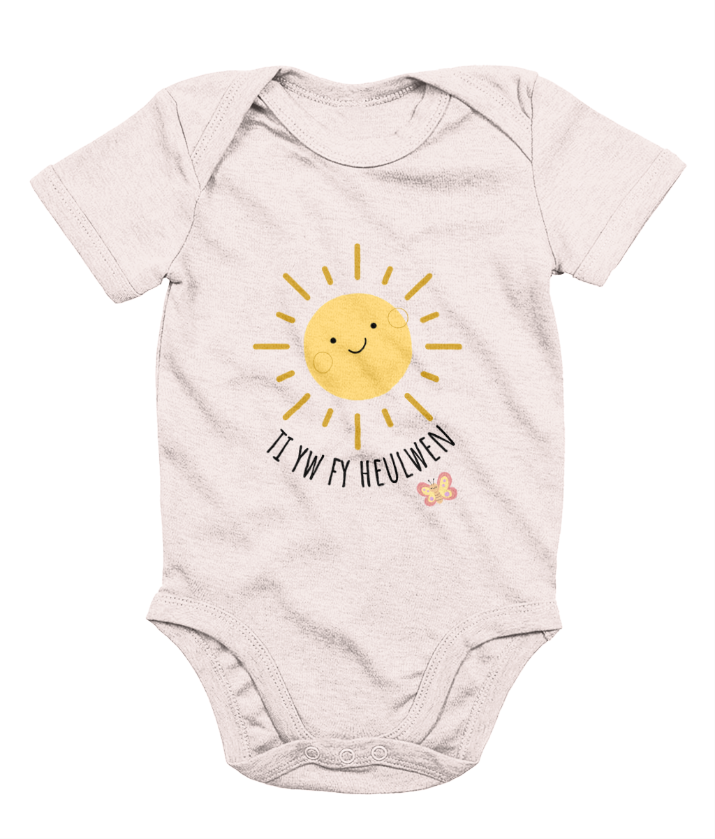 A baby pink babygrow with the text 'Ti yw fy heulwen' printed on the front, featuring a sun illustration and a small heart.
