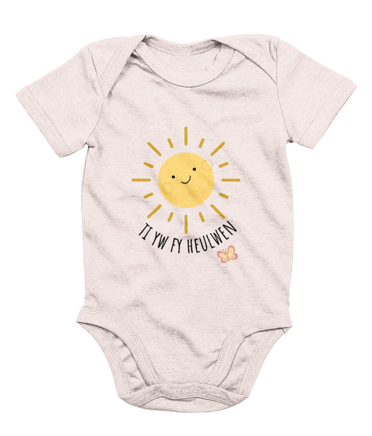 A baby pink babygrow with the text 'Ti yw fy heulwen' printed on the front, featuring a sun illustration and a small heart.