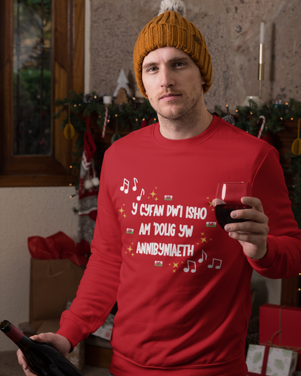 A person wearing a red Christmas sweatshirt with Welsh text and festive motifs, holding a drink and standing near holiday decorations.
