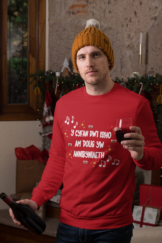 A person wearing a red Christmas sweatshirt with Welsh text and festive motifs, holding a drink and standing near holiday decorations.