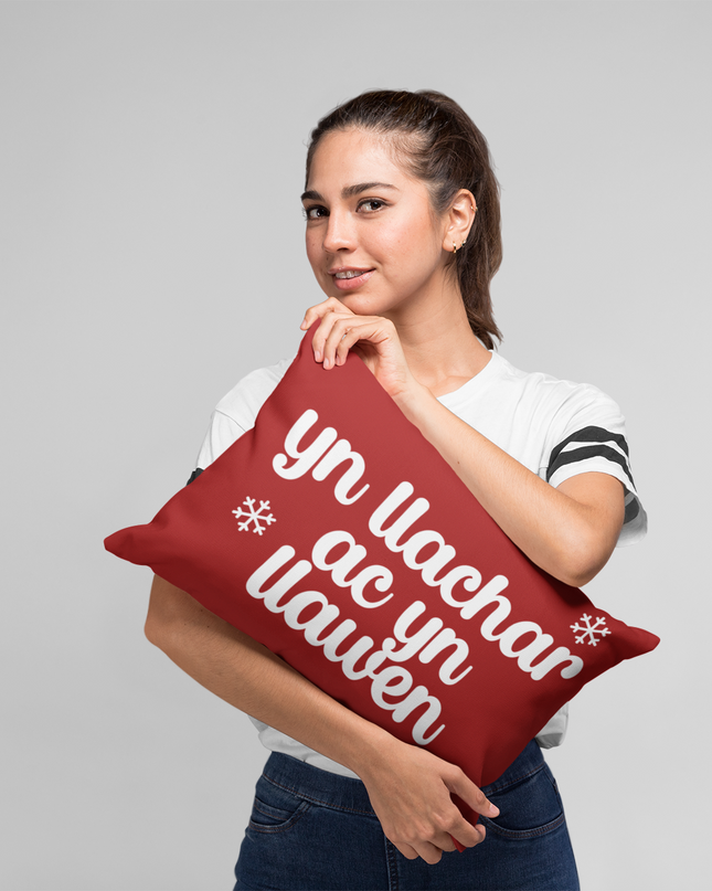 A person holding a red Christmas pillow with the text 'yn llachar ac yn llawen' printed in white, meaning 'Merry & Bright' in Welsh.